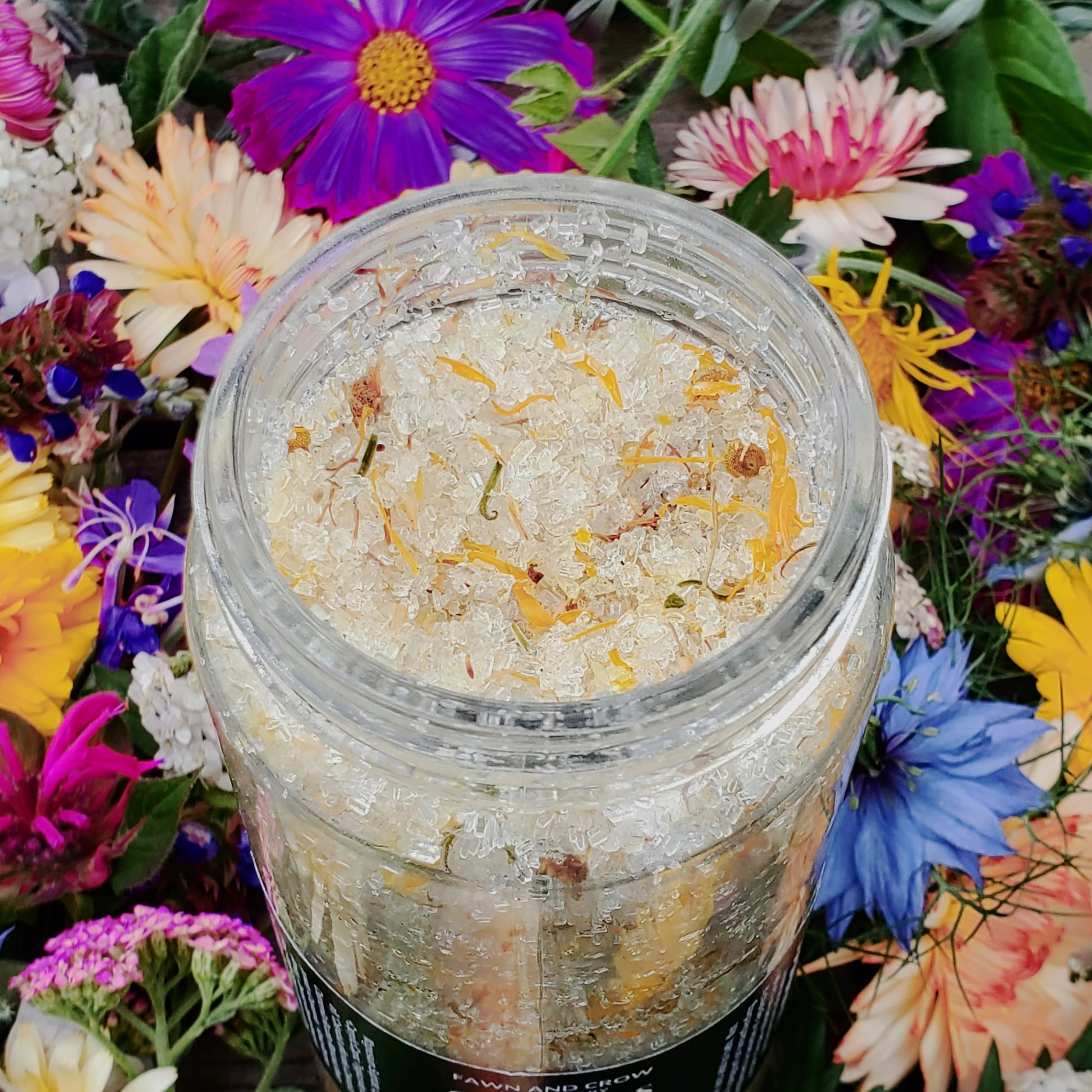 Botanical herbs and epsom salts by Fawn and Crow Apothecary in a clear jar, surrounded by colorful flowers