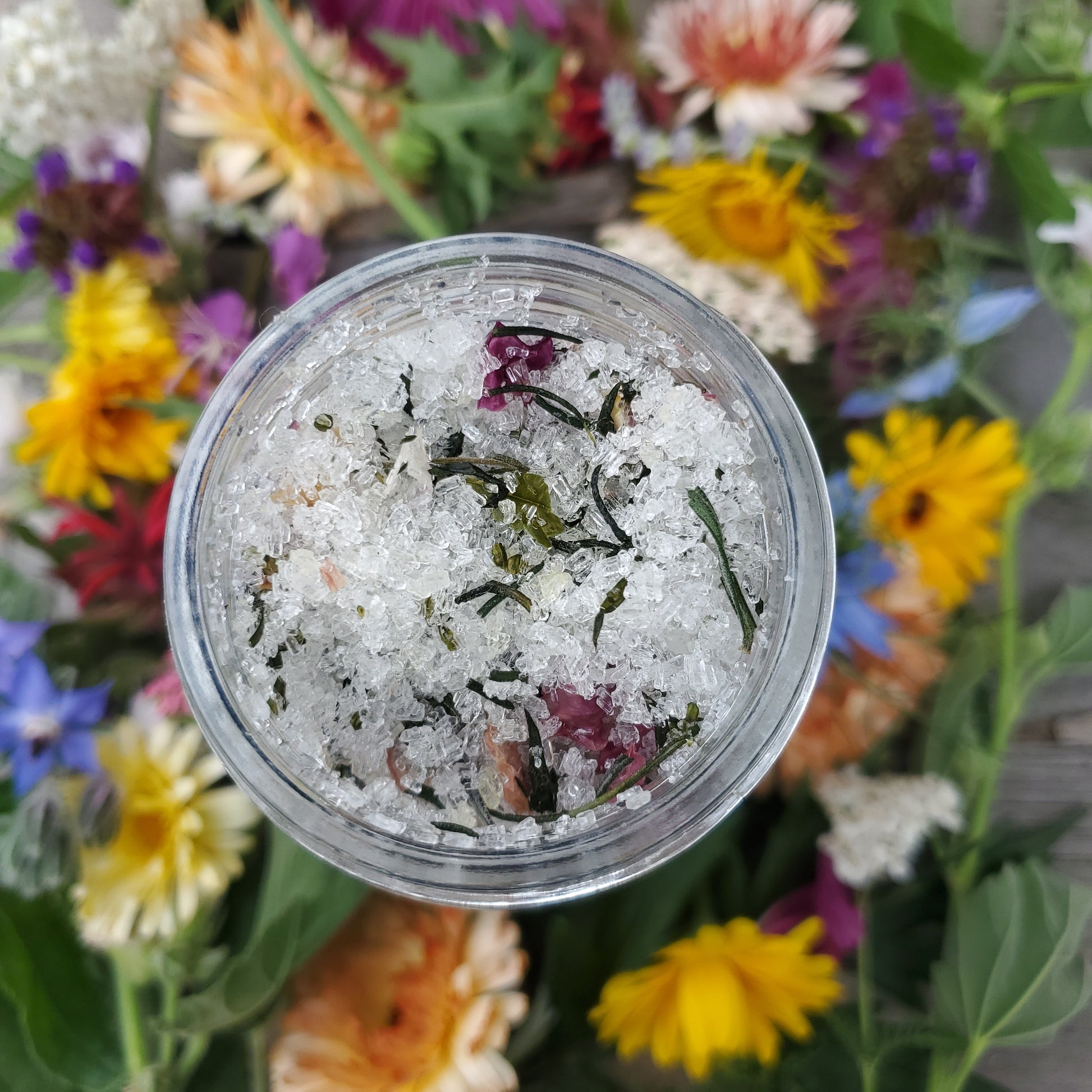 Epsom salts and herbs in a clear jar surrounded by colorful flowers.