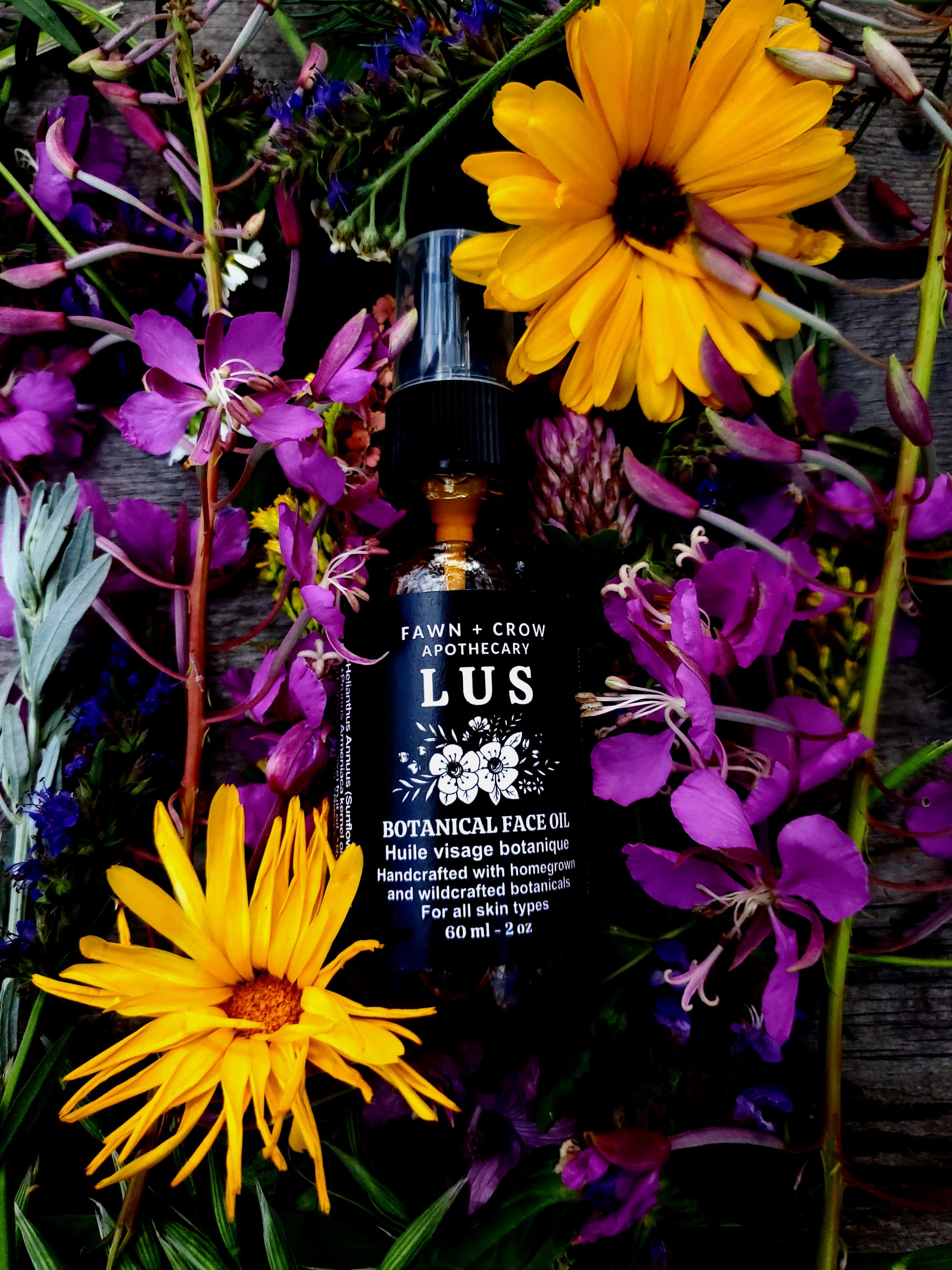 Bottle of Fawn & Crow Apothecary LUS Botanical Face Oil surrounded by yellow and purple flowers.