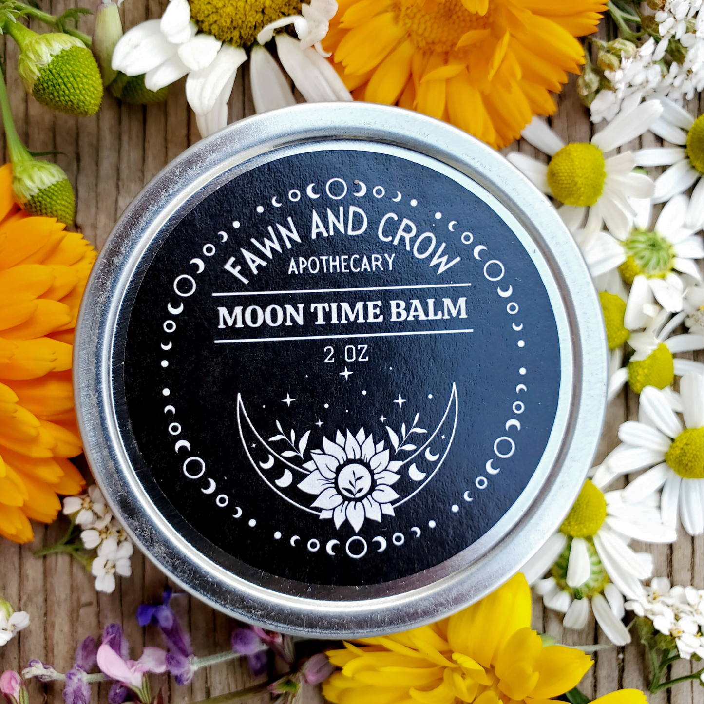 A tin container labeled 'Moon Time Balm' by Fawn and Crow Apothecary, surrounded by an array of wild flowers including Yarrow and other botanicals.