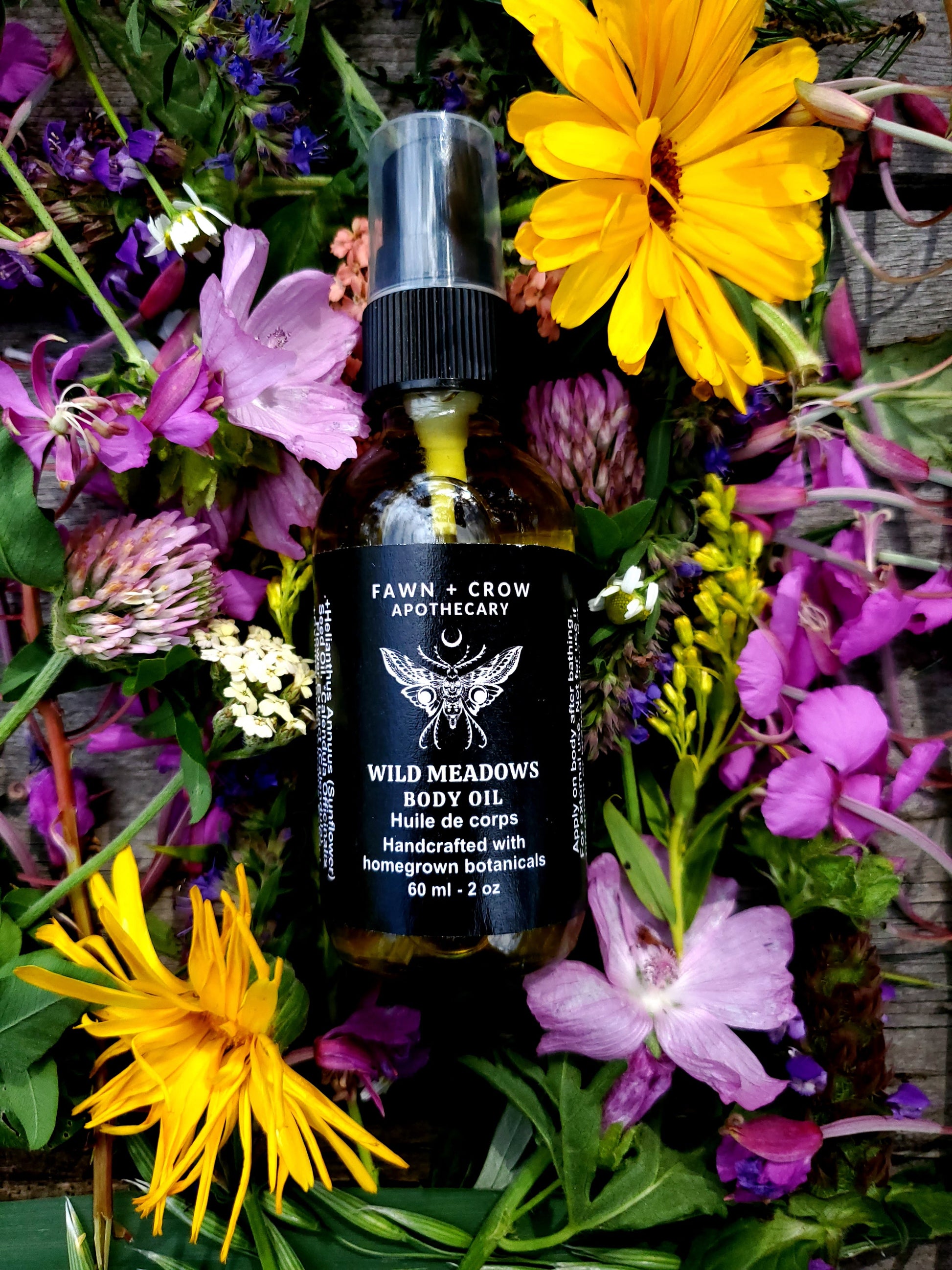 Bottle of Fawn + Crow Apothecary Wild Meadows Body Oil surrounded by wildflowers on a wooden surface.