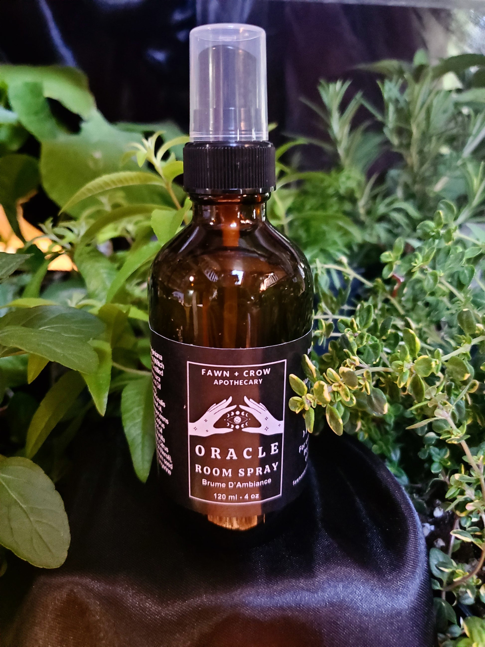 Brown glass bottle with spray nozzle labeled 'Oracle Room Spray' from Fawn & Crow Apothecary, set against a green leafy background.