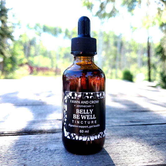 Bottle of 'Belly Be Well Tincture' by Fawn and Crow Apothecary on a wooden surface with a blurred natural background.